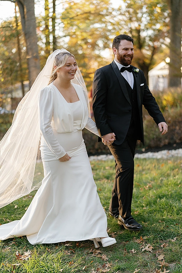 Wedding recessional bride and groom walking hand in hand across an outdoor lawn, her cathedral veil trailing behind, tent and autumn trees in daylight