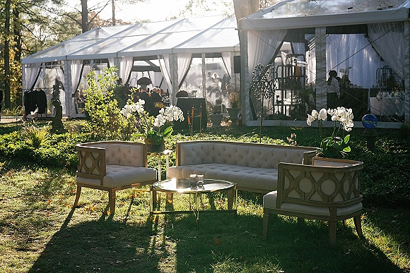 Wedding lounge area with outdoor wedding lounge sofas and chairs around a coffee table, white orchid centerpiece under a clear top tent on a sunny lawn