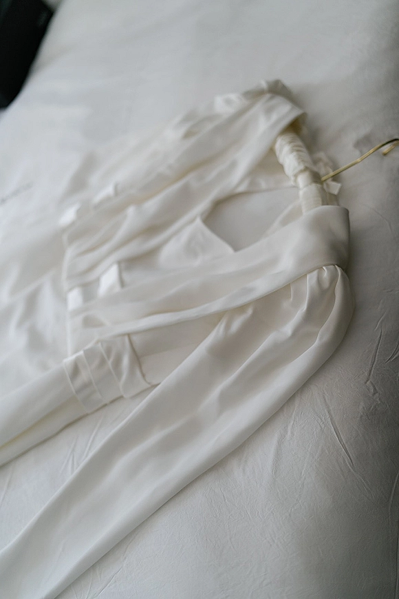 Wedding dress detail of a satin wedding dress with spaghetti straps and fabric-covered buttons on a padded hanger over white bedding