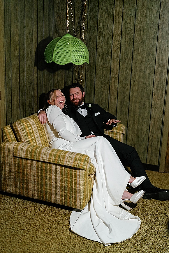 Couple portrait of bride and groom on couch, laughing as he holds her close in tuxedo and long sleeve gown against wood panel wall.