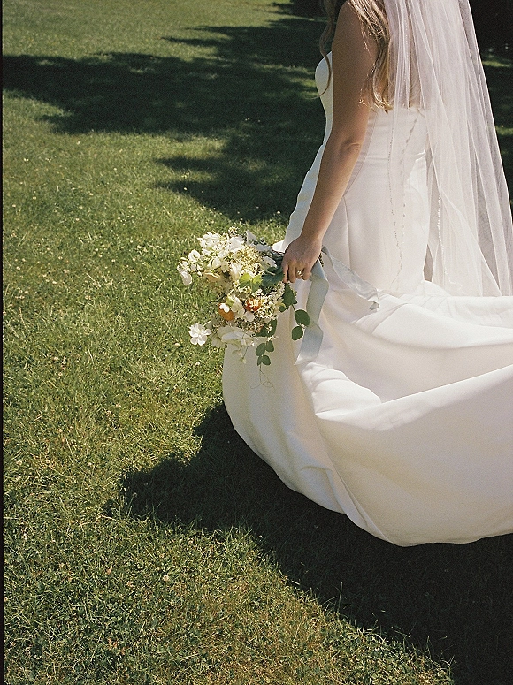 Bride portrait from behind holding bouquet, long bridal veil over strapless wedding dress, walking on grass lawn with tree shadows