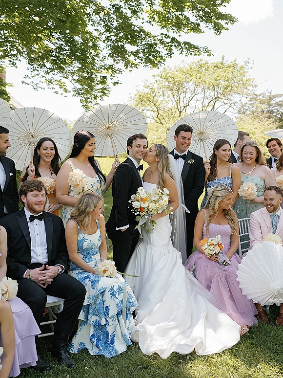 Wedding party photo of bride kissing groom as bridesmaids hold bouquets and paper parasols on a green lawn with trees in daylight