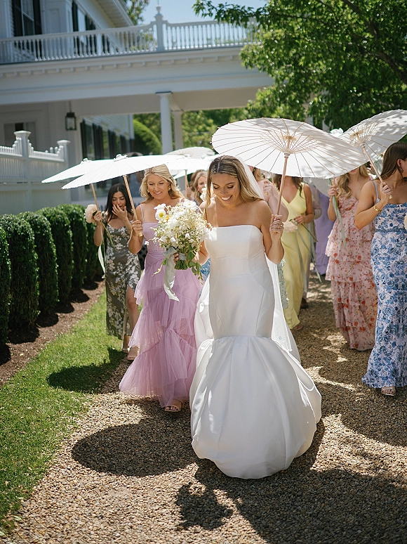 Bridal party processional with bride walking under a parasol, holding a bouquet, bridesmaids behind her on a gravel garden path