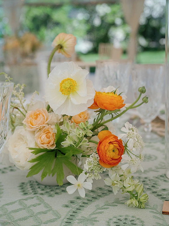Wedding centerpiece with a low floral centerpiece of white poppies, orange ranunculus, and peach roses in a ceramic bowl on a patterned tablecloth under a tented reception
