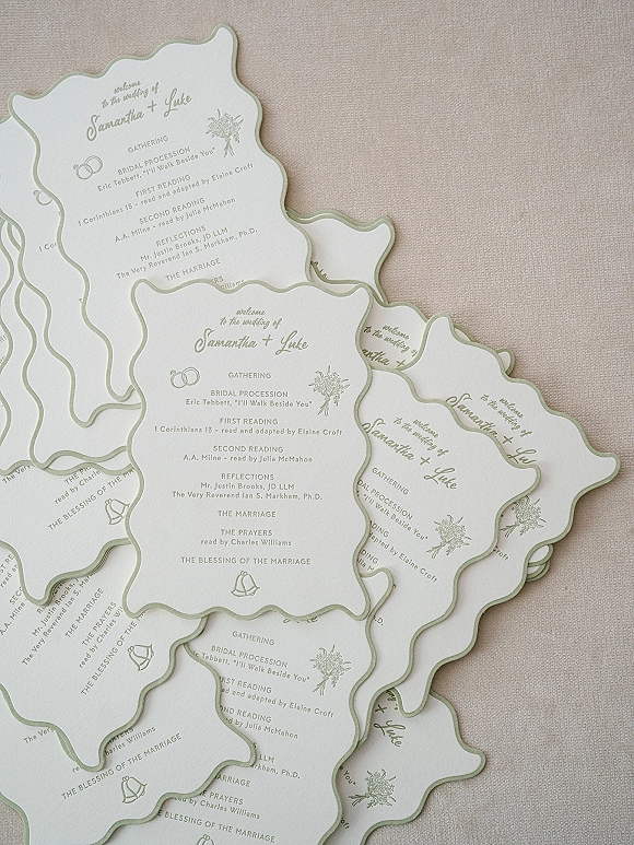 Wedding ceremony program with scalloped wedding program edge, sage green border and calligraphy, shown on a linen fabric flat lay