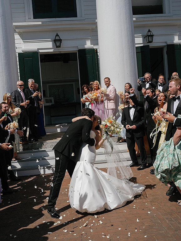 Wedding kiss portrait as groom dips bride in veil and strapless dress, guests cheer tossing petal confetti on courthouse steps