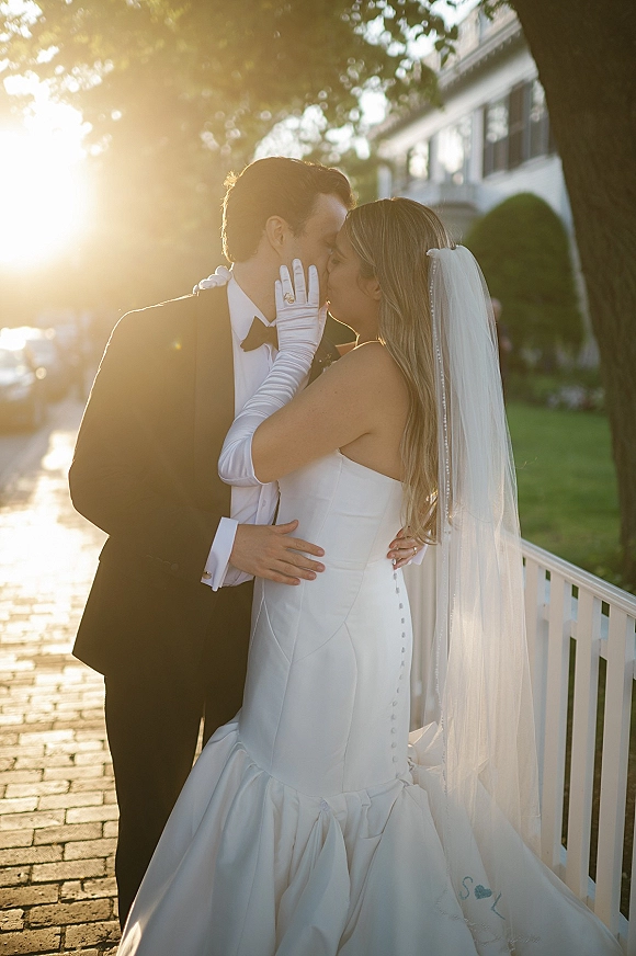 Wedding kiss portrait of bride and groom kissing at golden hour, her long gloves and veil framing the moment as sunlight flares near trees