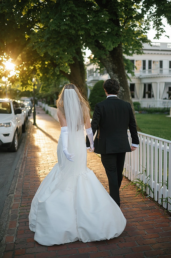 Newlywed couple holding hands, bride and groom walking away with a long veil on a brick sidewalk by a white picket fence at sunset
