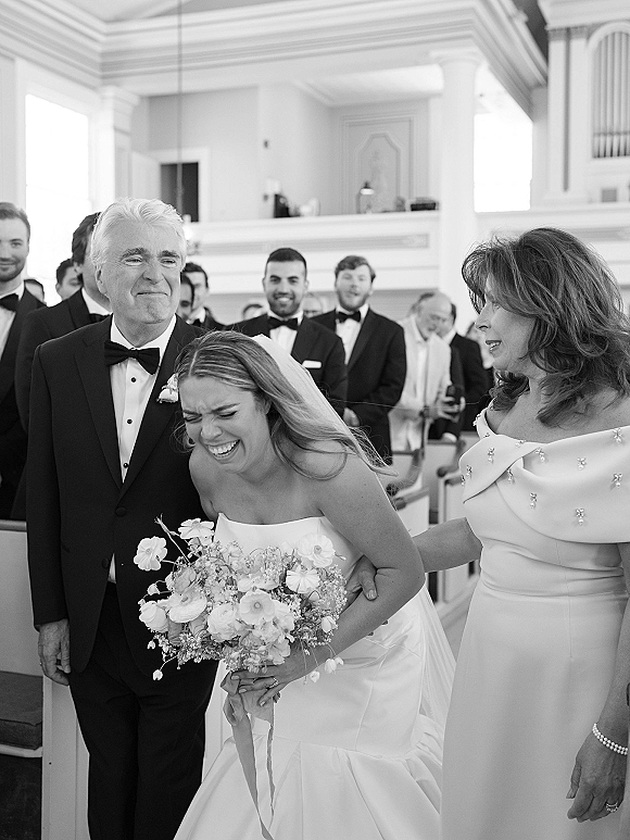 Bride with parents laughing in the church aisle, holding a bouquet with long ribbon wrap as wedding guests look on from wooden pews