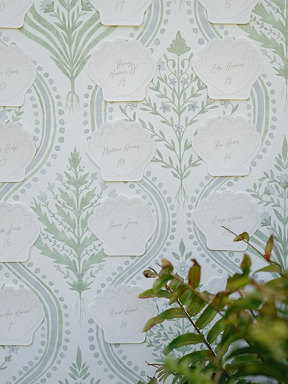 Seating chart wedding seating chart display with scalloped calligraphy place cards arranged on a green and white botanical pattern wall