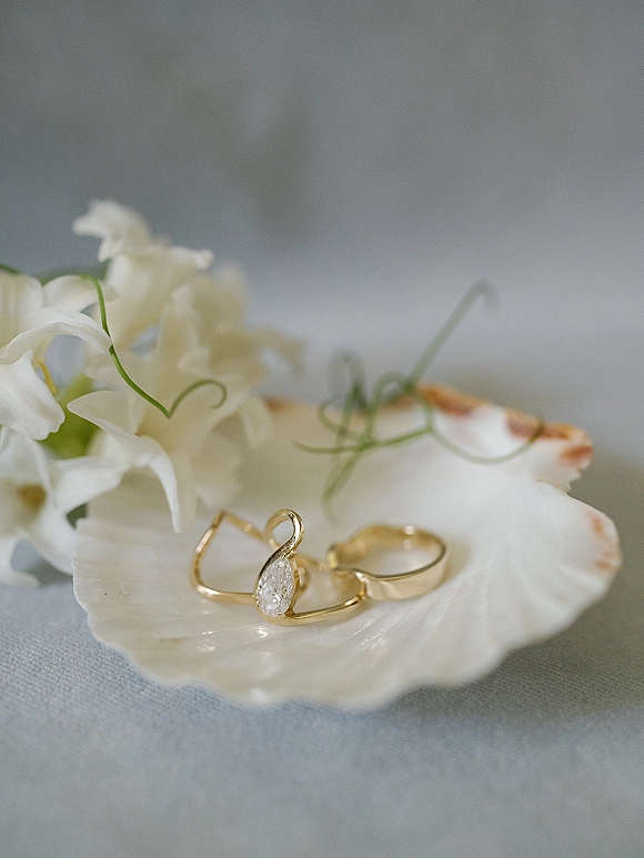 Wedding rings arranged in a wedding ring flatlay on a shell dish with a diamond engagement ring and white flowers on linen backdrop