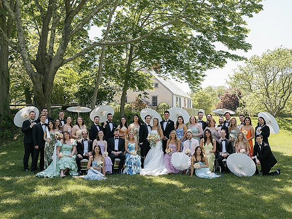 Wedding party portrait with bride and groom with wedding party, bridesmaids in pastel dresses and groomsmen in tuxedos on lawn, white parasols
