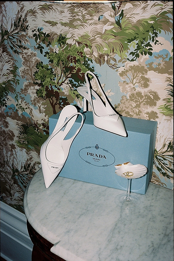 Bridal shoes, white slingback heels on a marble table beside a ring dish with wedding rings, set against botanical wallpaper corner