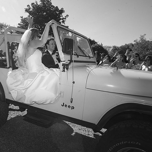 Wedding exit as bride waves from a jeep wedding exit, veil flowing in vintage convertible while groom drives past cheering guests on gravel road
