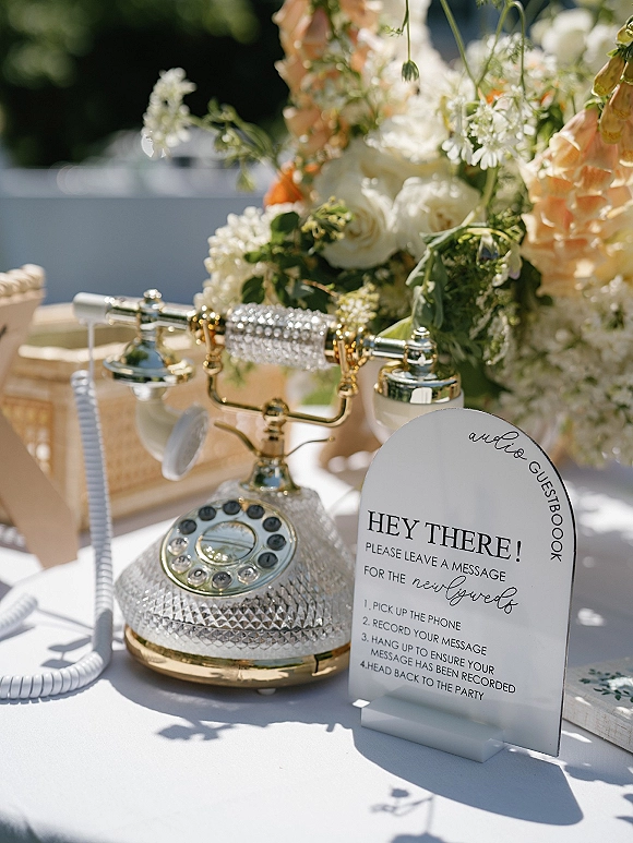 Audio guestbook wedding audio guestbook with a vintage telephone and guestbook sign beside white and peach flowers on a white tablecloth outdoors