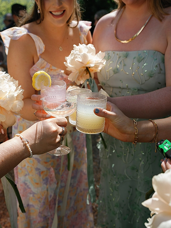Wedding cocktail toast as bridesmaids clink coupe and textured glasses with lemon garnish, gold jewelry, and peonies in sunlit greenery