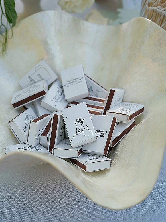 Wedding matchbooks and custom wedding matchboxes with illustrated covers arranged on a tray dish beside greenery and a white flower