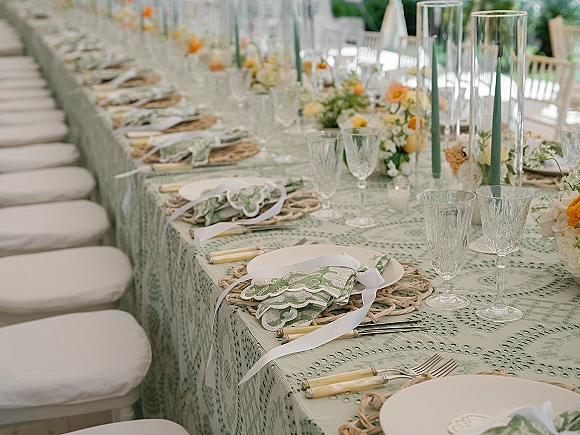 Reception tablescape with long banquet table setup on green patterned tablecloth, rattan chargers, gold flatware, crystal goblets, taper candles outdoors