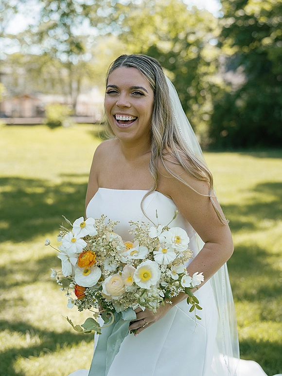 Bridal portrait of a laughing bride holding a white and yellow bouquet, wearing a strapless dress and veil on a sunlit lawn with trees