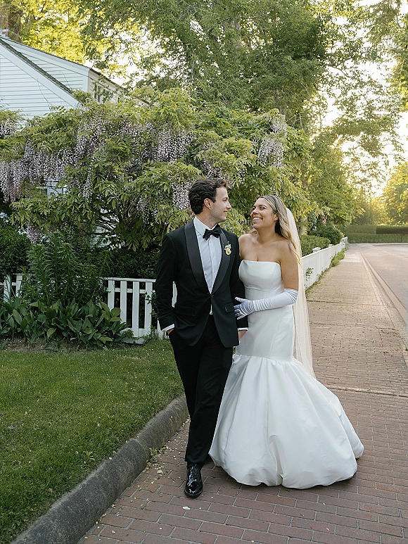 Couple portrait of bride in strapless dress and veil with opera gloves and groom in black tuxedo, strolling by a white picket fence garden walkway