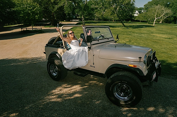 Wedding getaway car jeep wedding exit with bride in a strapless dress and sunglasses in a convertible on a sunny park road