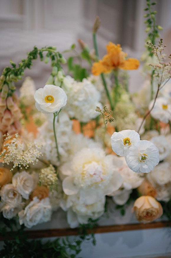 Wedding floral arrangement with white blooms and greenery, dotted with orange buds in a floral foam box against a light stone wall and window frame