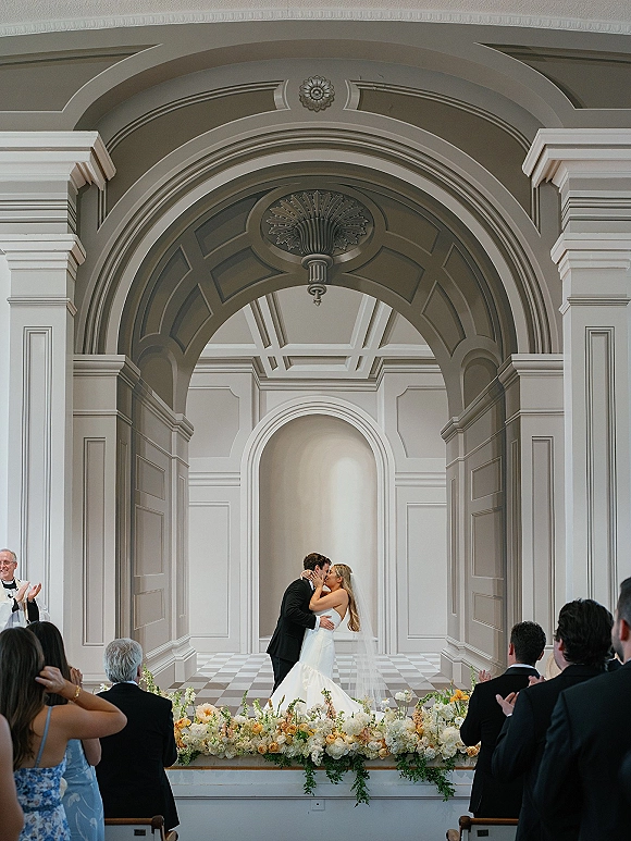Ceremony kiss as bride in strapless wedding dress and veil kisses groom in black suit at altar under arched doorway indoors