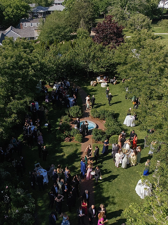 Outdoor wedding cocktail hour with aerial wedding reception view, white linen cocktail tables and lounge seating around a garden fountain on a brick pathway