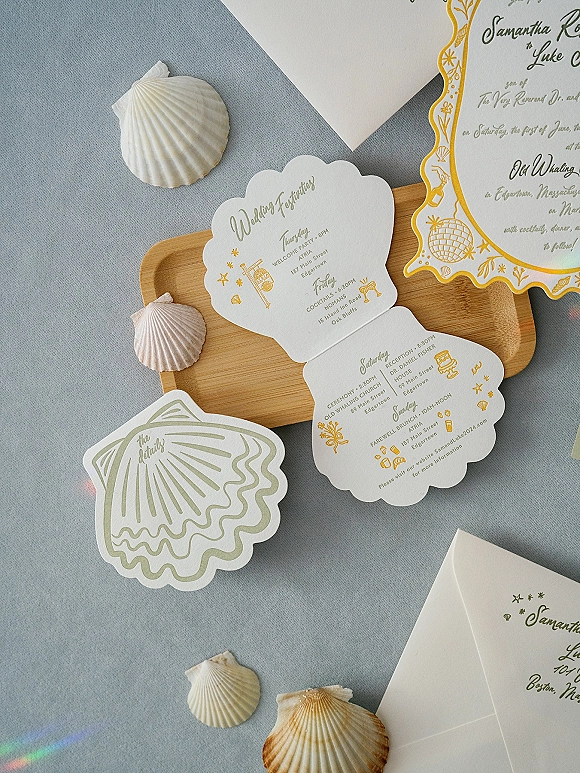 Invitation suite of scallop-shaped beach wedding invitations with gold and green ink, envelope and seashells on blue fabric surface
