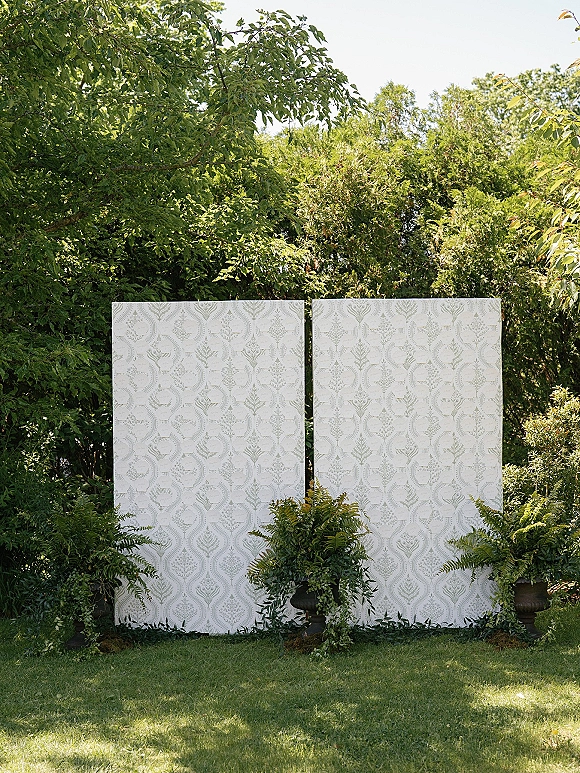 Ceremony backdrop with patterned panels framed by lush greenery and potted ferns, set on a grassy garden lawn in daylight