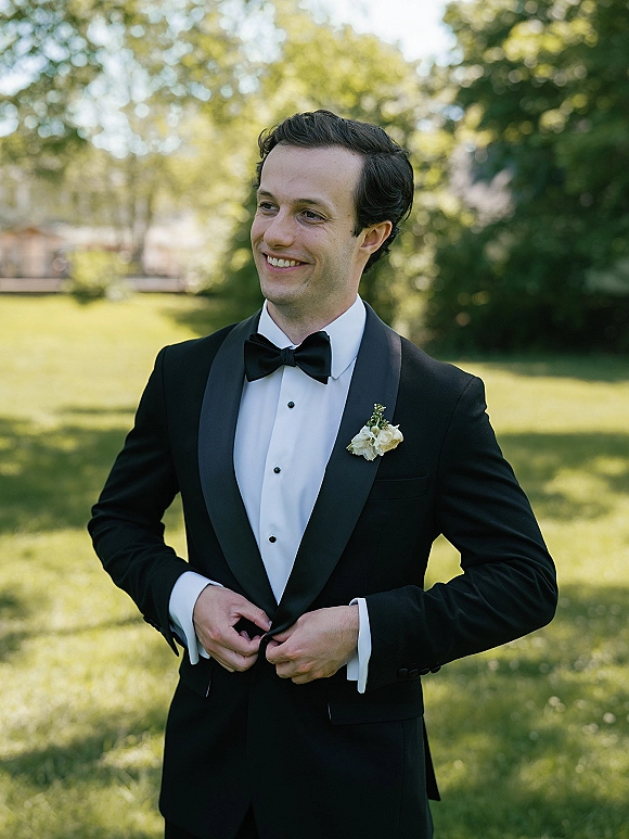 Groom portrait in a black tuxedo with bow tie and boutonniere, smiling in sunlight on a green lawn with trees behind