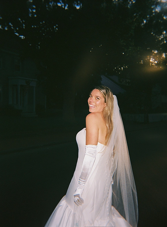 Bridal portrait of a smiling bride looking over shoulder in a strapless dress with veil and opera gloves on a tree-lined walkway at night