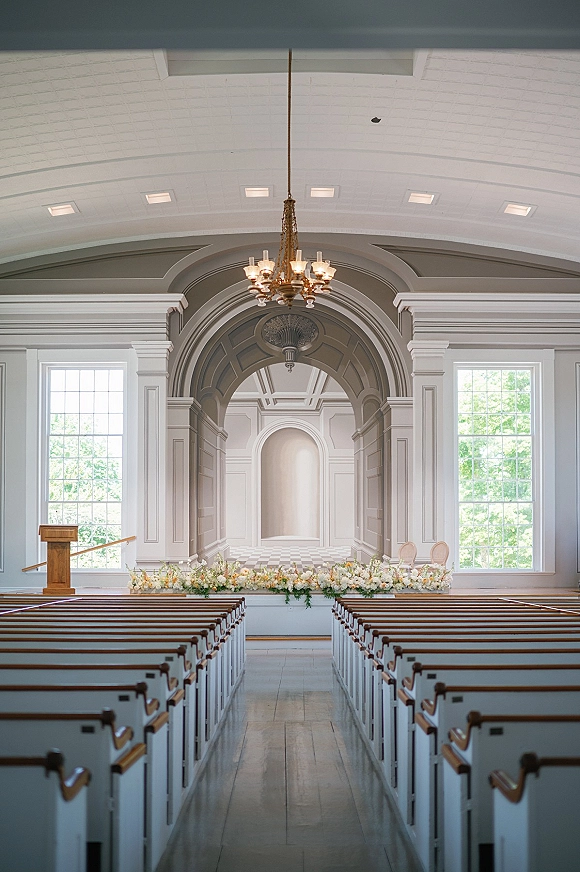 Ceremony setup in a bright white chapel with gold chandelier, floral altar arrangement, wooden podium, and chairs lining the aisle