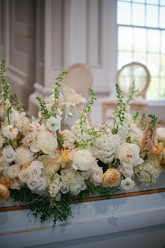 Wedding floral arrangement with white roses and peach garden blooms spilling along a ledge, set against white paneled walls and a large window