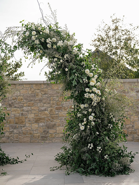 Wedding floral arch with an asymmetrical wedding arch design of greenery and white blooms, set against a stone wall patio with trees and sky