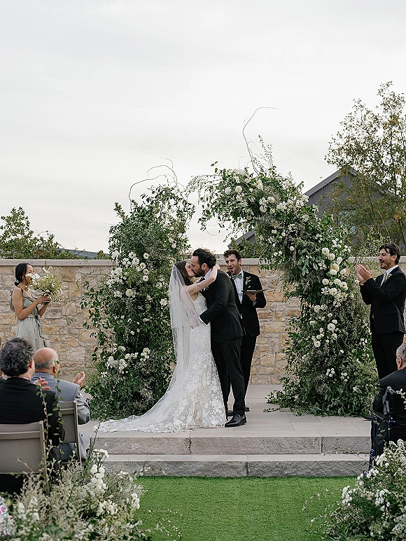 Ceremony kiss as bride in lace dress and veil kisses groom in black tux beneath a white flower and greenery arch in a courtyard