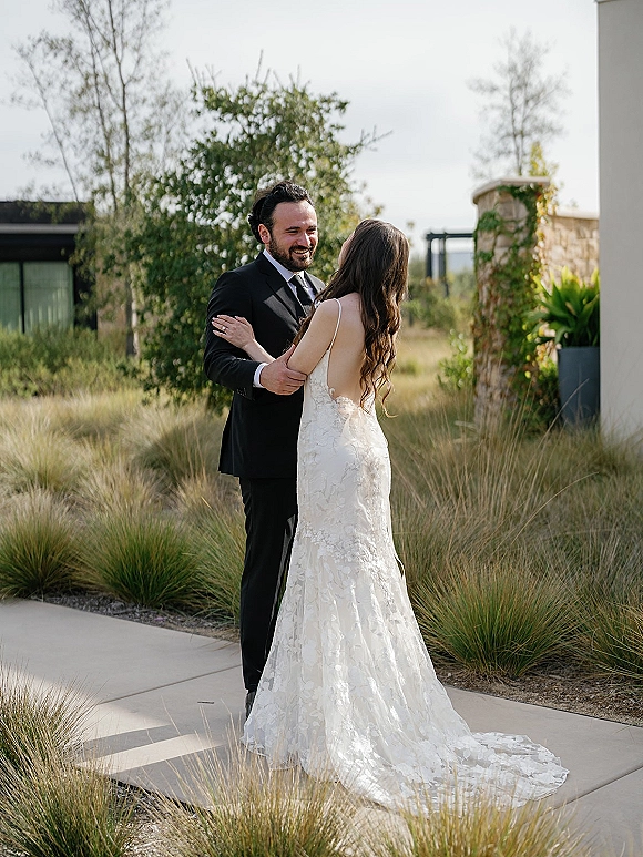 Couple portrait of bride and groom embracing, her lace wedding dress with low back and train, in a landscaped garden by a stone wall