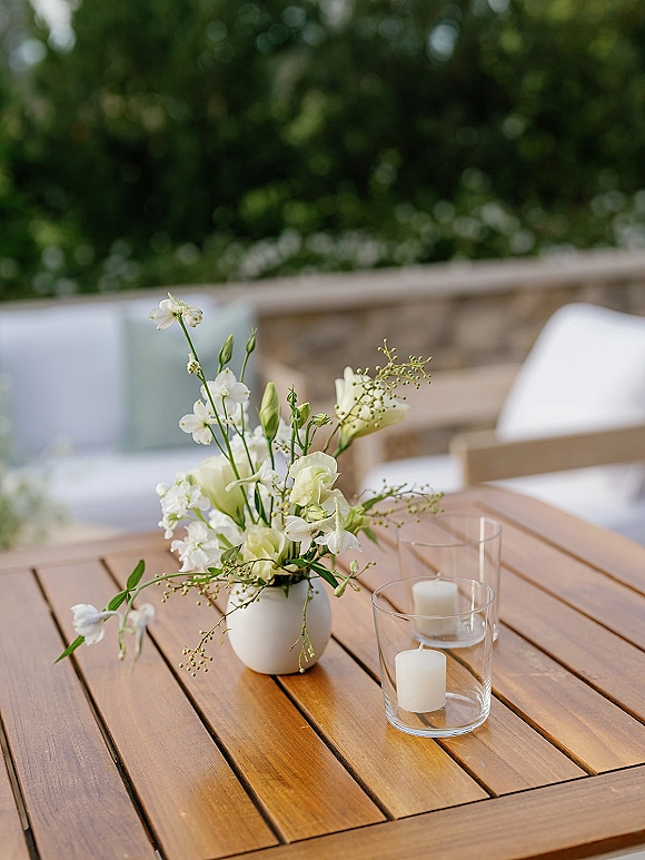 Wedding table centerpiece with white floral arrangement in a ceramic vase, glass candle holders and pillar candles on a wooden table outdoors