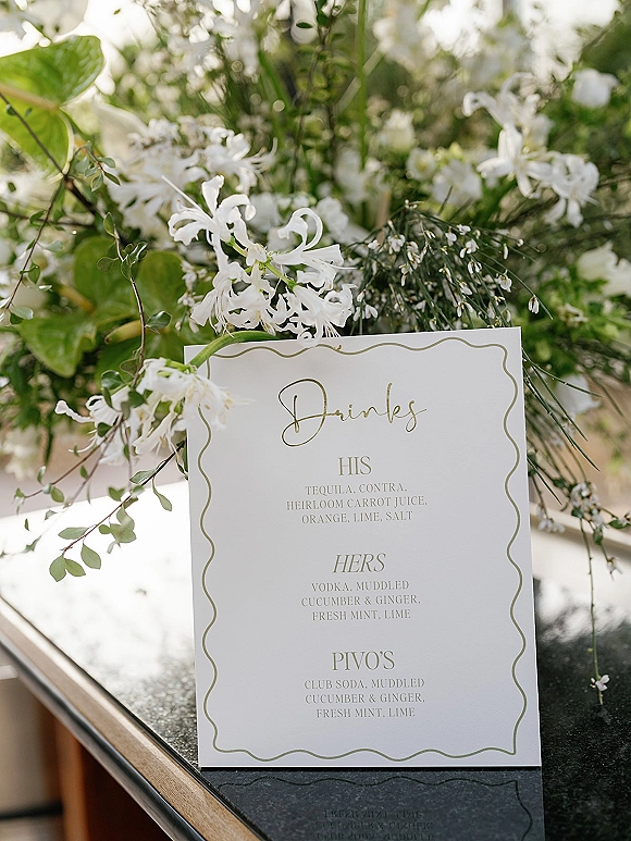 Wedding drinks menu card with scalloped border and calligraphy beside white floral arrangement and greenery on a black stone bar countertop