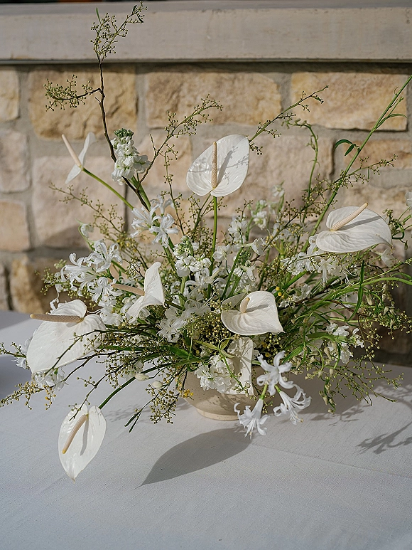 Wedding centerpiece with white anthurium centerpiece blooms and lush greenery in a ceramic bowl vase on a tablecloth against a stone wall