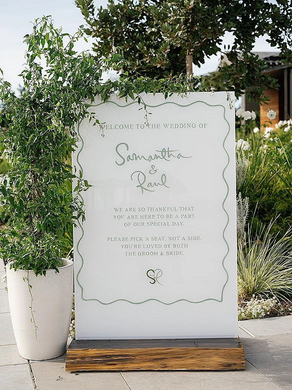 Wedding welcome sign with sage green lettering on a white scalloped board, set on a wood base beside a potted vine in a garden entry