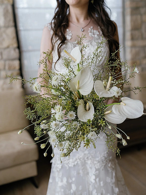 Bridal bouquet with white anthurium wedding bouquet blooms and greenery, held against a lace bodice and pendant necklace indoors by a stone wall