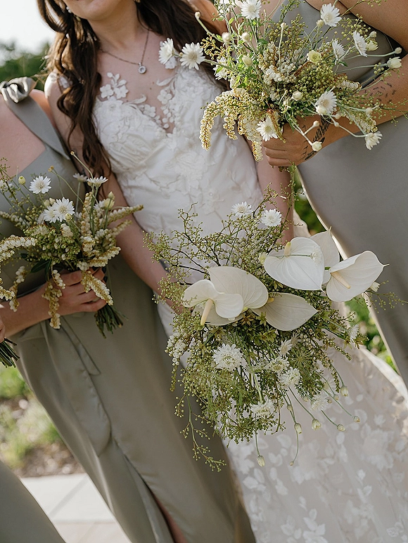Bridesmaid bouquets held against sage green bridesmaid dresses, featuring white anthurium, daisies, and greenery in outdoor greenery setting