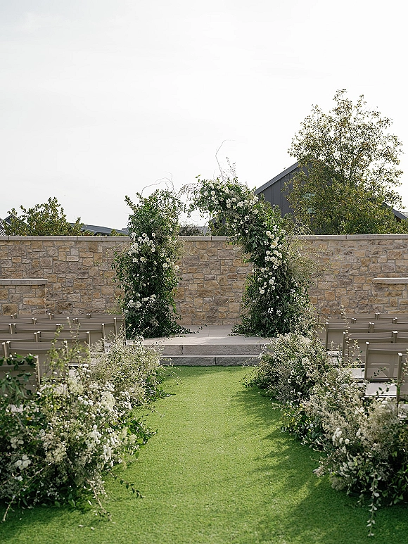 Ceremony aisle design with outdoor wedding aisle leading to a round floral arch of white flowers and greenery on a grassy lawn by a stone wall