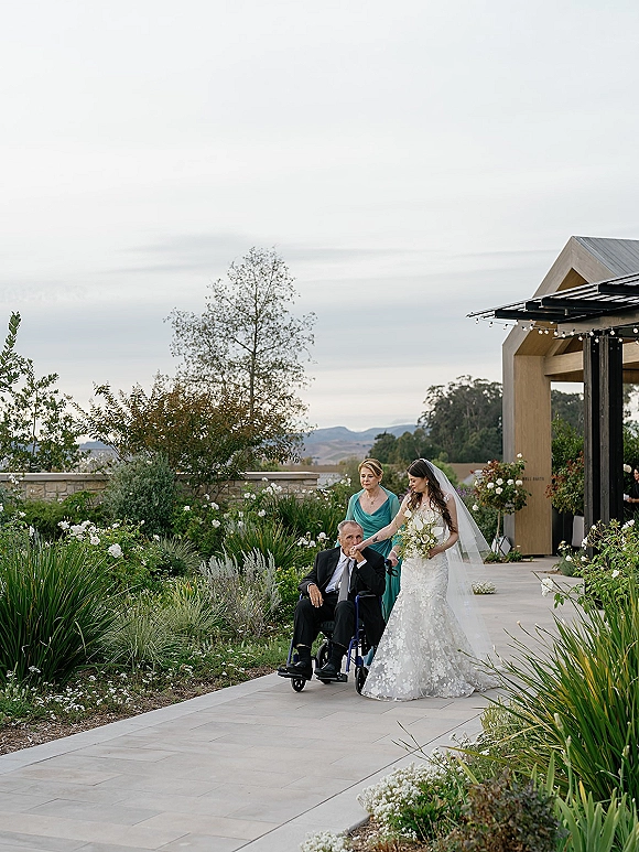 Bride with father walking beside his wheelchair, holding a white and green bouquet, veil trailing along a garden walkway with string lights