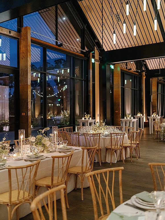 Reception tablescape with round wedding reception tables set in white linens, taper candles and white florals in a glass-walled room at dusk