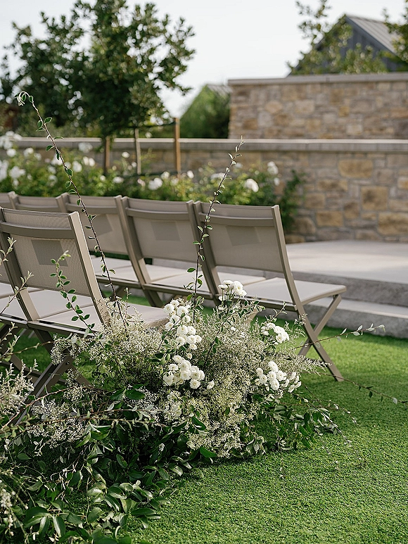 Ceremony seating with outdoor wedding ceremony chairs in neat rows, aisle lined with white flowers and greenery on a grass lawn by a stone wall
