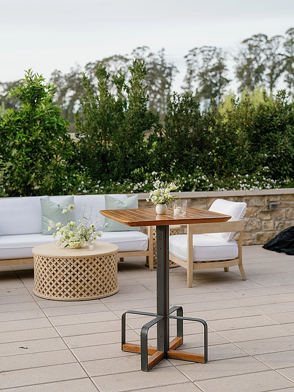 Wedding lounge setup with a white outdoor sofa, chairs, wood tables, and floral accents on a stone patio by hedges and trees