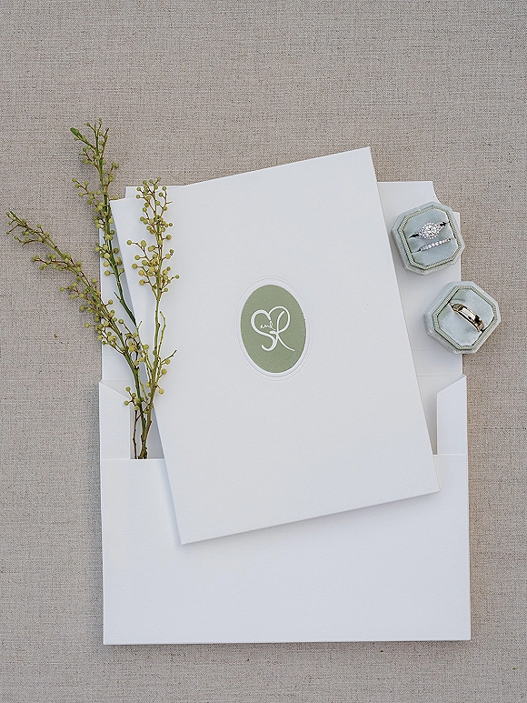 Invitation suite wedding invitations flat lay on linen fabric with envelopes, monogram sticker, greenery sprig, ring boxes, and rings