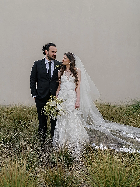 Couple portrait of bride and groom portrait looking at each other, bride holding bouquet with long veil trailing beside tall grass wall